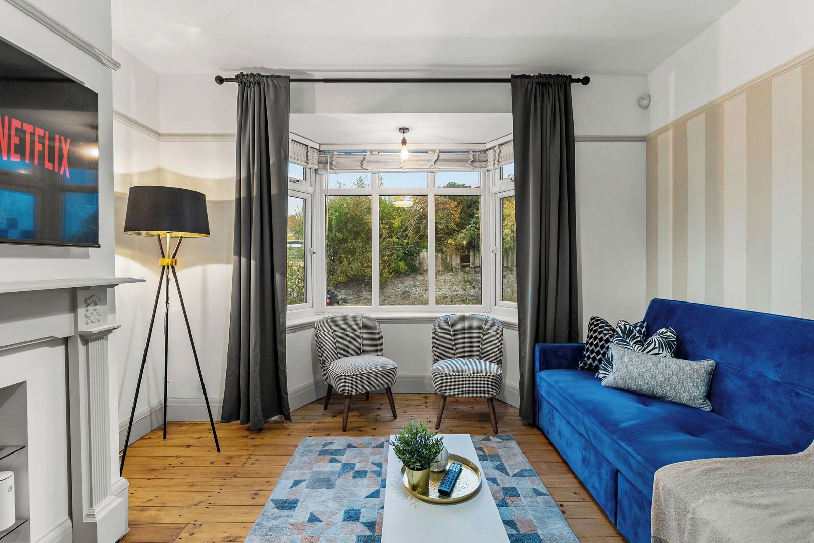 A living room with a blue sofa, two armchairs, a floor lamp, a TV, and a window with curtains.