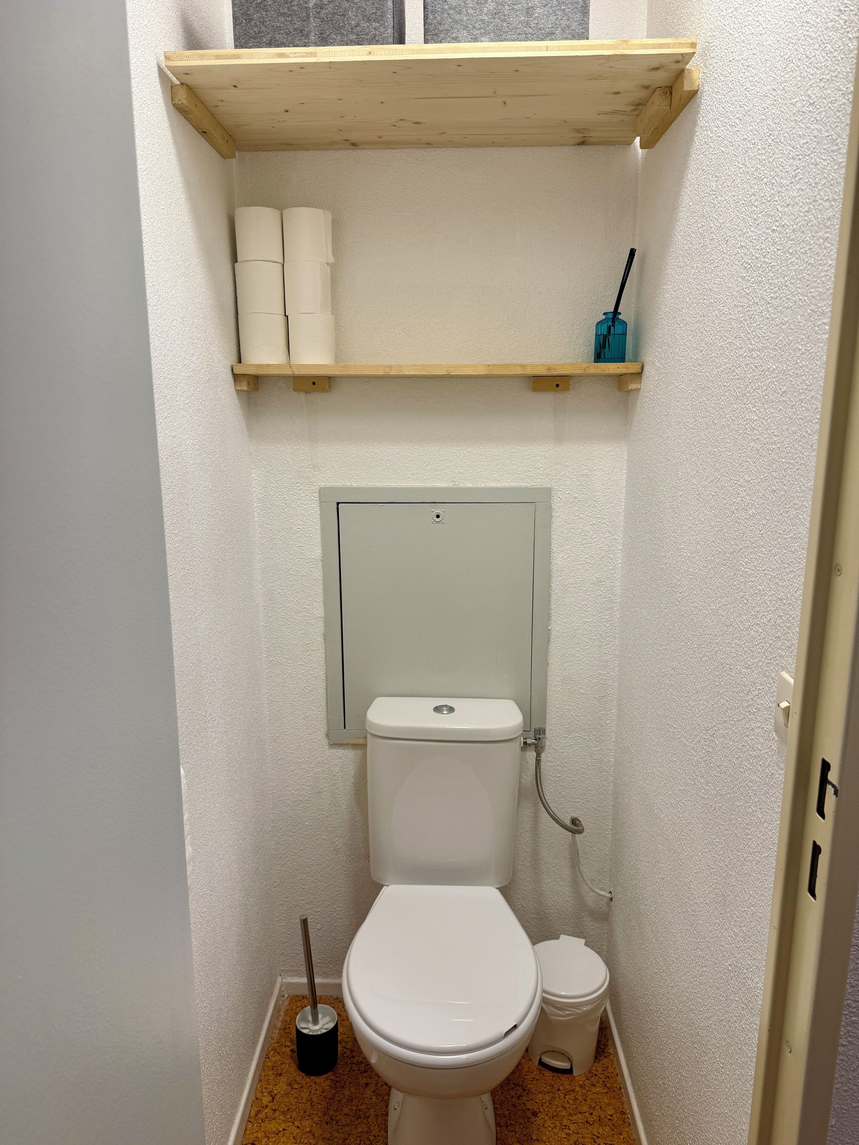 Ein kleines Badezimmer mit Toilette, einem Regal über der Toilette mit Toilettenpapier und Bürste sowie einem Spiegel an der Wand.