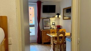 A narrow hallway leading to a room with a wooden table, chairs, a TV mounted on the wall, and a window with red curtains.