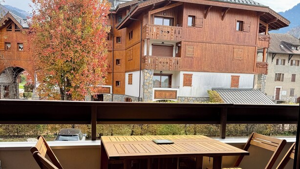 A wooden balcony with a table and chairs, overlooking a traditional building with balconies and a tree with autumn leaves.