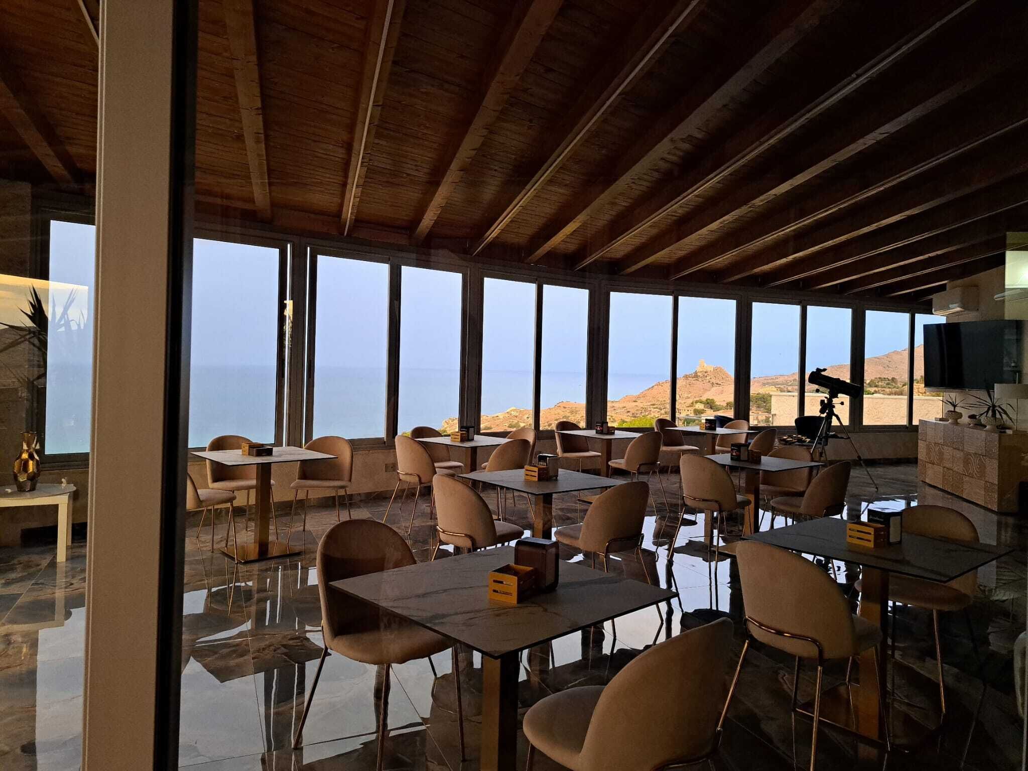 Un restaurante con vistas al mar y a las colinas, con vigas de madera, ventanas amplias y mobiliario moderno.