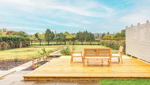 Una terraza de madera con banco y sillas, rodeada por un jardín cercado con árboles y arbustos.