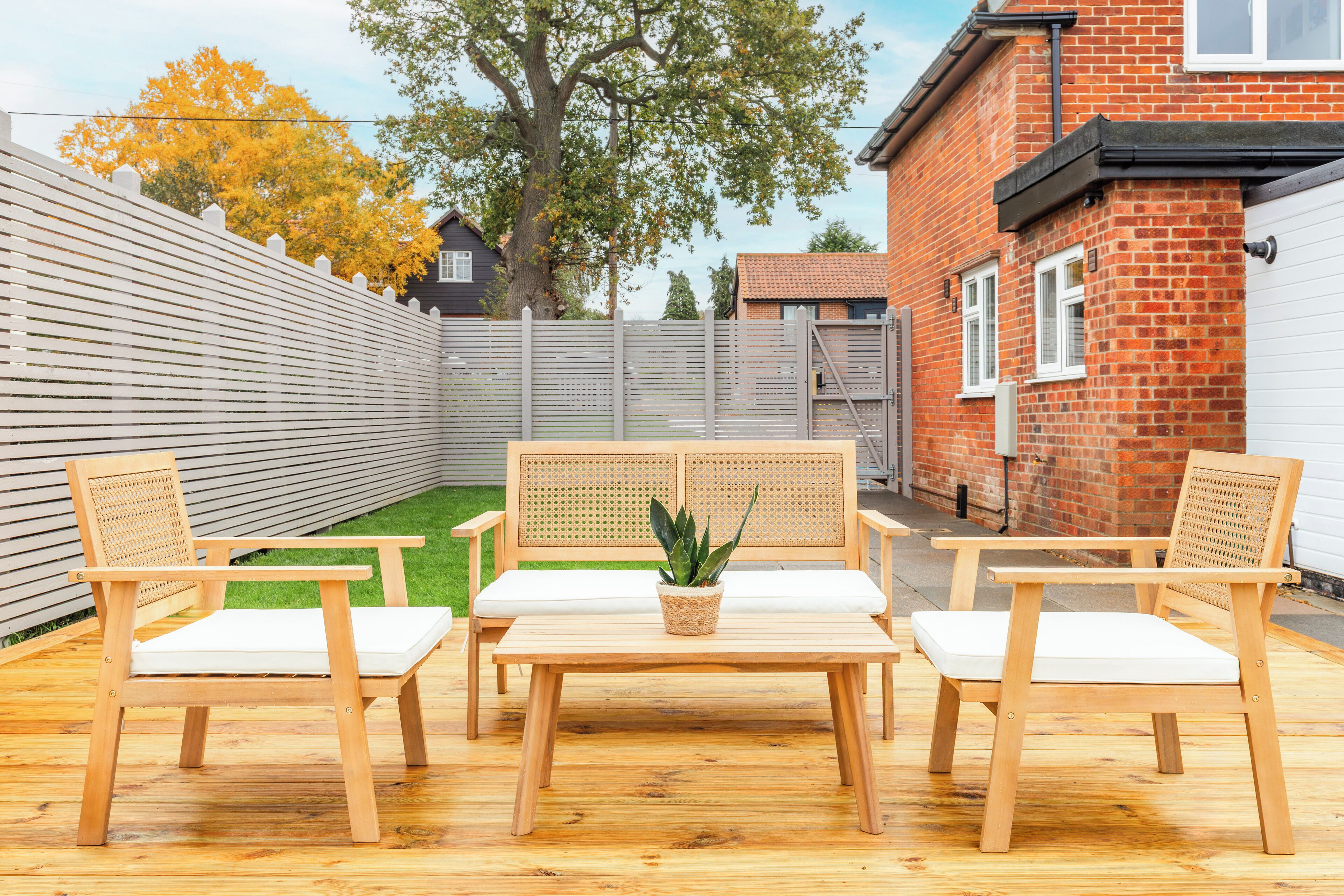 A wooden outdoor seating area with a table and chairs, a potted plant, and a white fence.