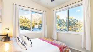 A bedroom with two large windows, a bed with a colorful blanket, a bedside lamp, and a view of trees and mountains.