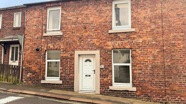 Une maison à deux étages, construite en briques, avec une porte et des fenêtres blanches, une cheminée bien visible et une rue devant.