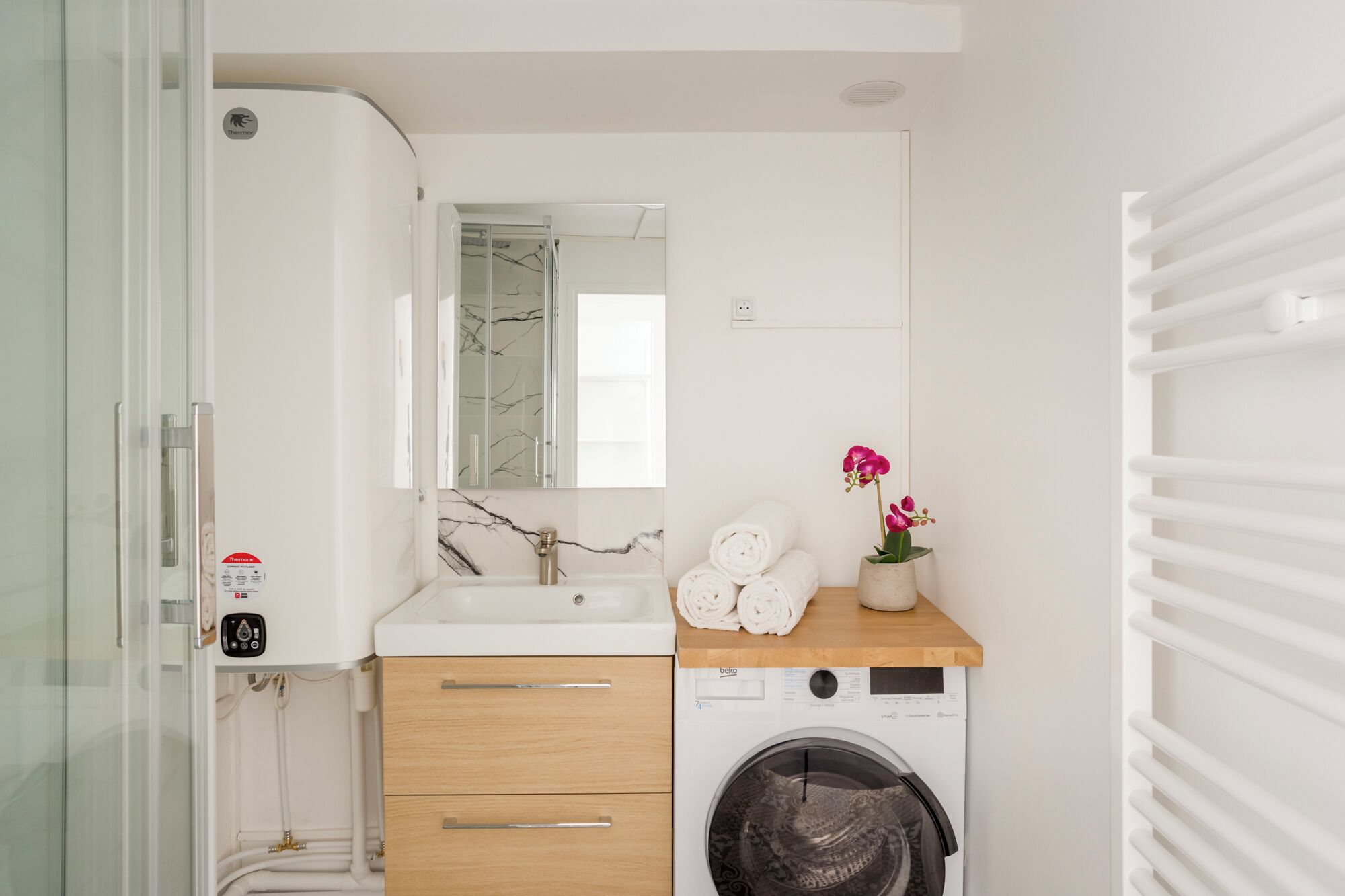 Un baño con un lavabo blanco, espejo y lavadora. Hay toallas y una orquídea en maceta sobre el mostrador.