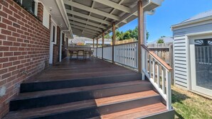 A covered outdoor deck with steps leading to a patio area, a wooden fence, and a building with a window. - Gwendoline Cottage, Pets, Main St. Station (Croydon)