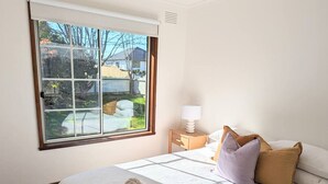 A neatly made bed with a rolled towel, a nightstand with a lamp, and a window with a view of a backyard. - Gwendoline Cottage, Pets, Main St. Station (Croydon)