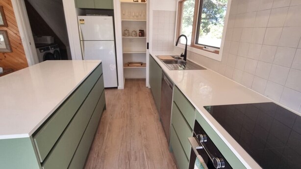 Een kleine keuken met groene kastjes, een wit aanrecht, een ingebouwde oven en een raam voor natuurlijk licht.