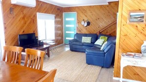 Een gezellige woonkamer met een blauwe bank, houten wanden, een eettafel met stoelen en een televisie.