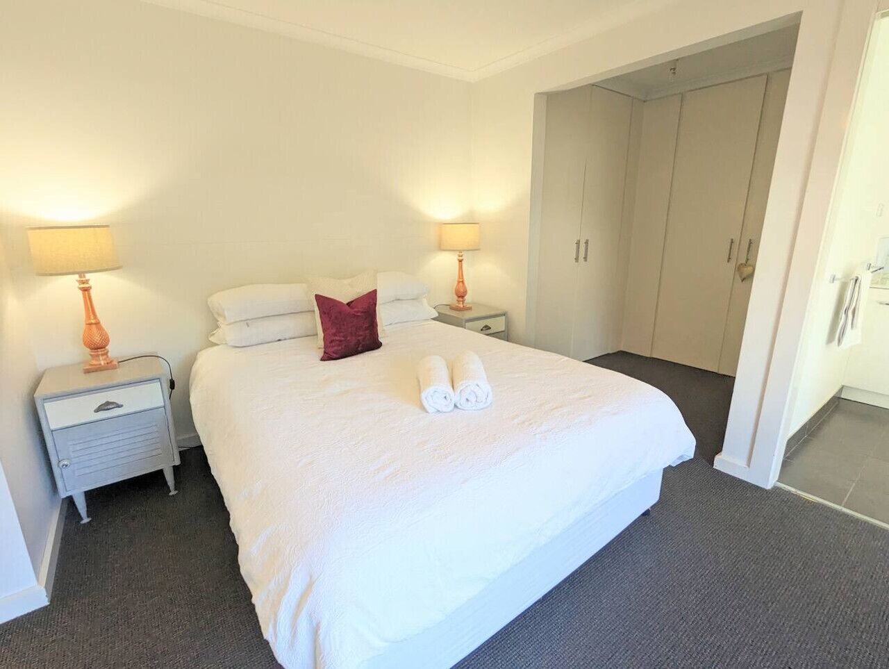 Hotelkamer met een groot bed, twee nachtkastjes met lampen, een rood decoratief kussen en een set opgerolde handdoeken op het bed.