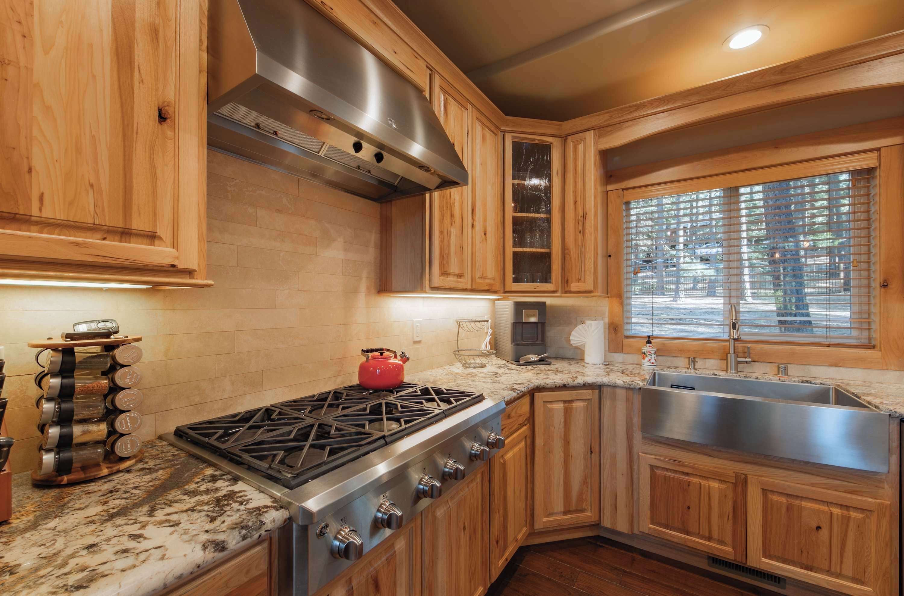 Une cuisine avec des meubles de rangement en bois, un évier en acier inoxydable, une cuisinière à gaz et une fenêtre avec des stores.