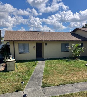 A single-story building with a brown roof, a front door, and a small front yard with a pathway leading to the door.