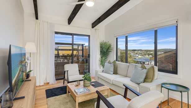 A modern living room with a sofa, coffee table, and a TV. There are large windows offering a view of the outside.