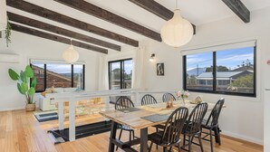 A dining area with a wooden table and chairs, a view of the outside through large windows, and a hanging light fixture.