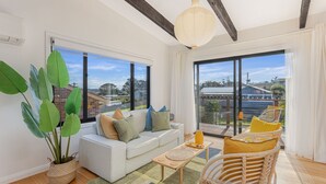 A modern living room with a sofa, a coffee table, and a wicker chair. There are large windows with a view of the neighborhood.