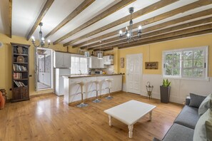 Un salón amplio con suelo de madera, una mesa de centro blanca, un sofá gris y una cocina con armarios blancos y una isla central.