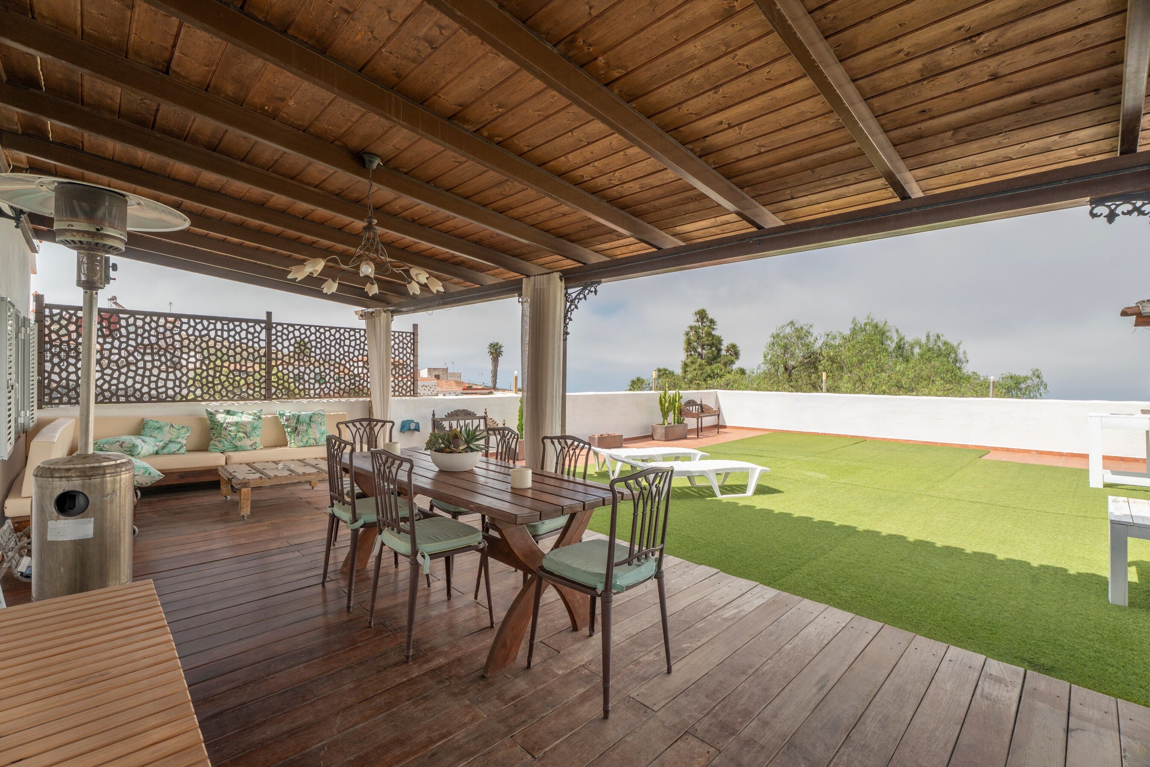 Una terrazza esterna con pavimento in legno, un tavolo da pranzo con sedie, un divano con cuscini e vista su un prato verde e alberi.