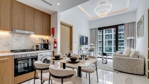 A modern kitchen with a stove, oven, and microwave. There is a dining area with a table set for four, chairs, and a sofa. The room has large windows and a view of buildings.