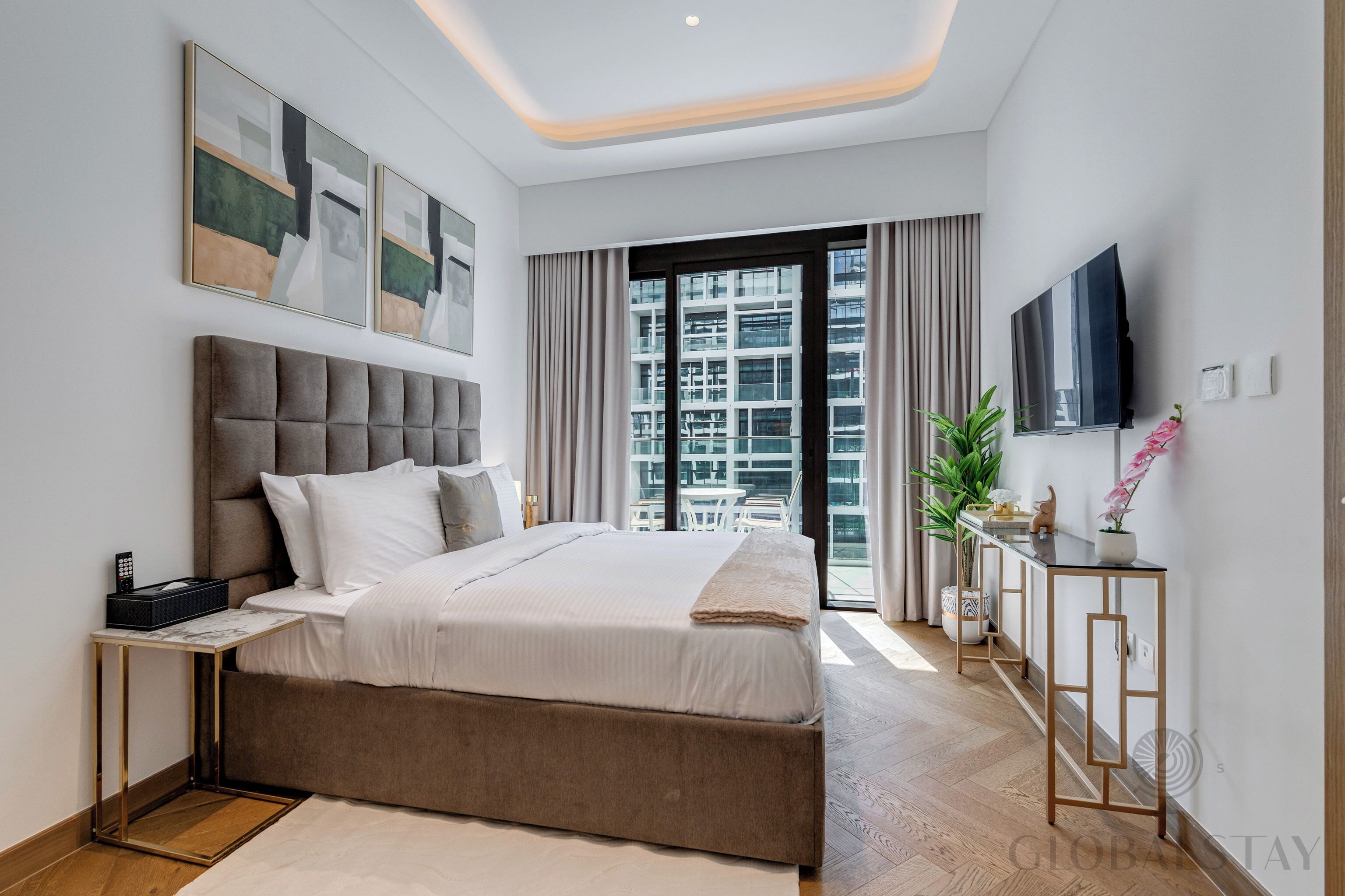 A modern bedroom with a large bed, a bedside table, a TV, and a view of the city through the window.