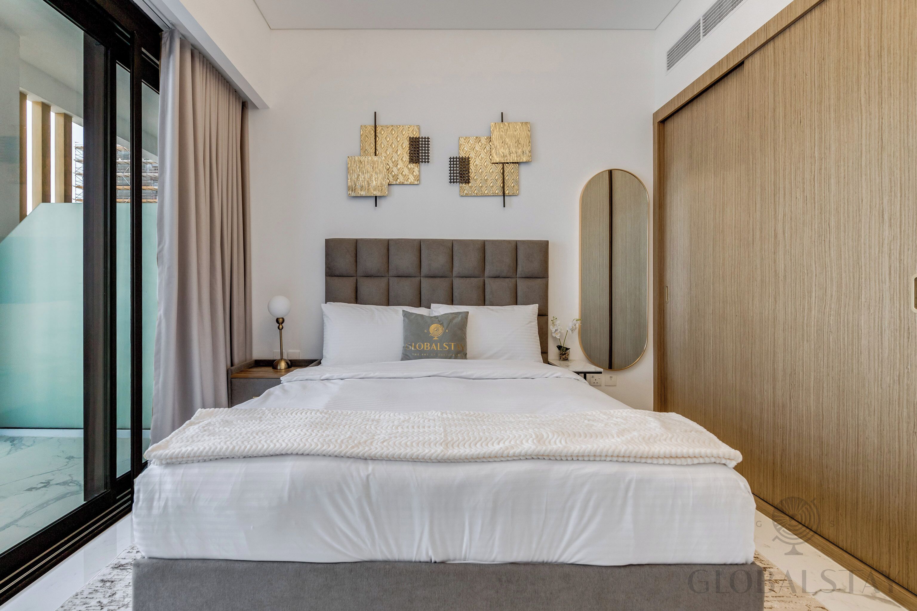Una habitación de hotel moderna con una cama grande, vistas a través de un ventanal, un armario de madera y un elemento decorativo en la pared.
