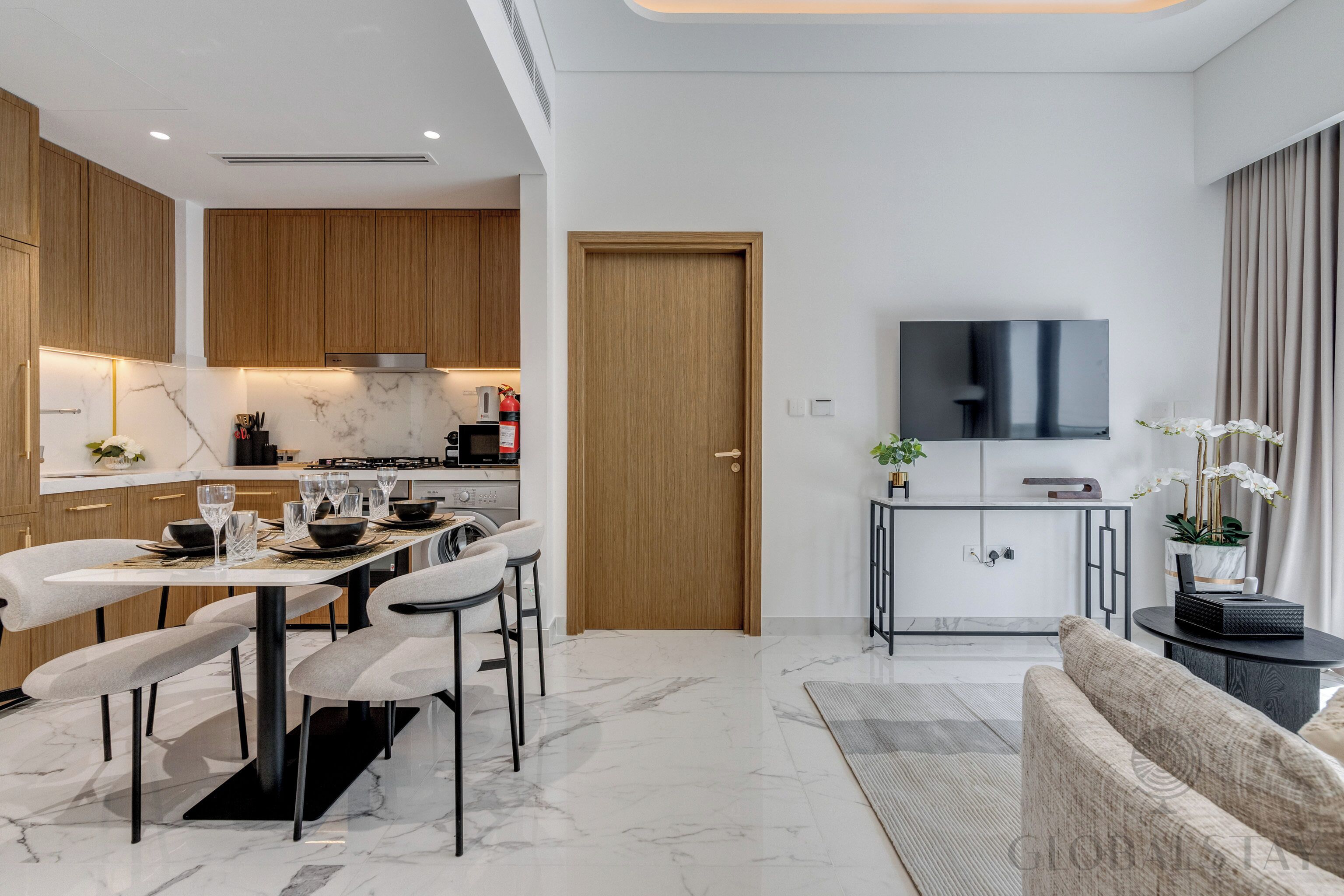A modern kitchen with wooden cabinets, a dining area with a marble table and chairs, a TV mounted on the wall, and a sofa in the living area.
