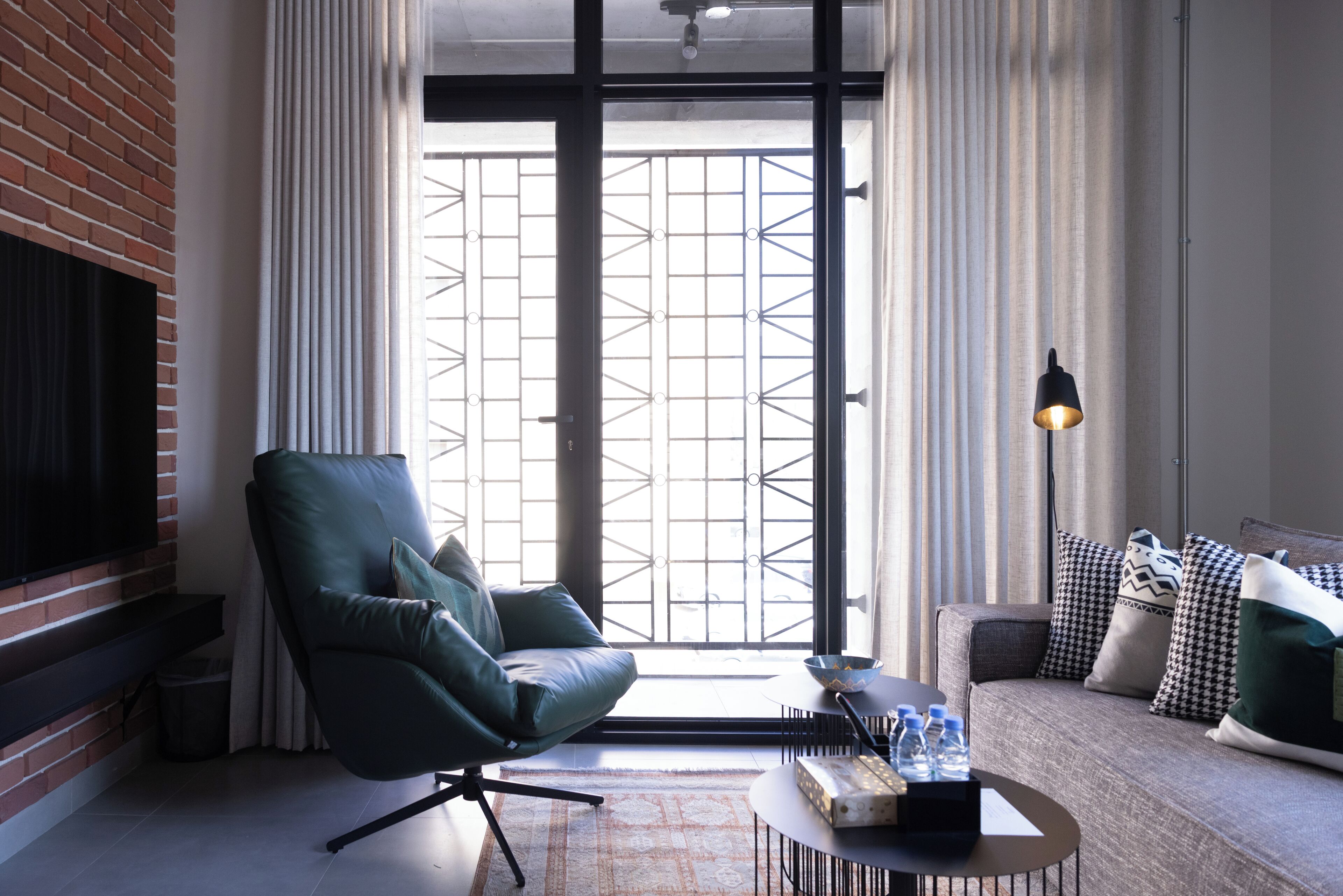 A modern living room with a leather armchair, a sofa with patterned cushions, a coffee table with a tray of bottled water, and a view through a large window with a metal grid.