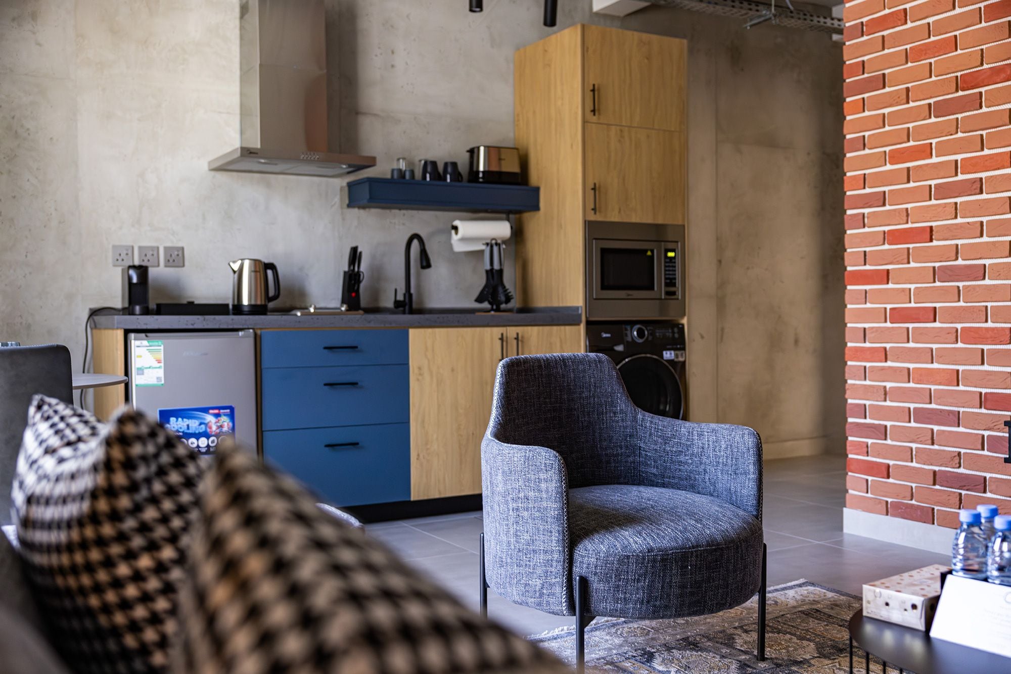 A modern kitchen with a microwave, kettle, and coffee maker. There is a blue chair and a sofa in the living area.