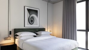 A modern hotel room with a large bed, a patterned rug, and a view of the city through large windows. - C LIVING 3 - ALMALQA (Riyadh)