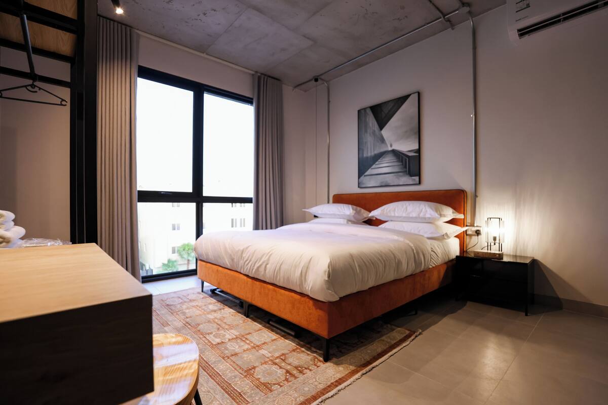 A modern bedroom with a large bed, a nightstand, a desk, and a window with curtains.