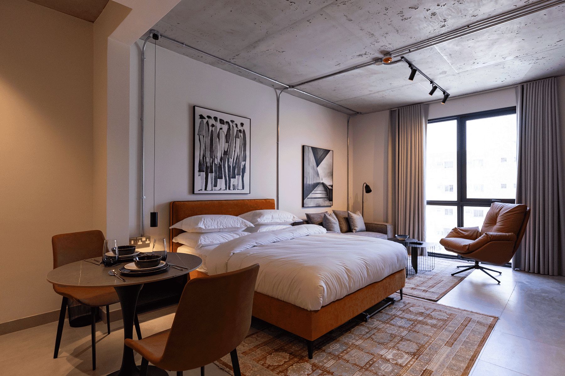 A modern hotel room with a large bed, a dining table, a chair, and a view of the city through large windows.