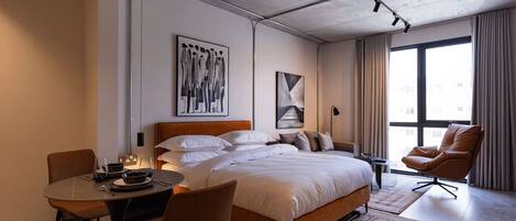 A modern hotel room with a large bed, a dining table, a chair, and a view of the city through large windows.