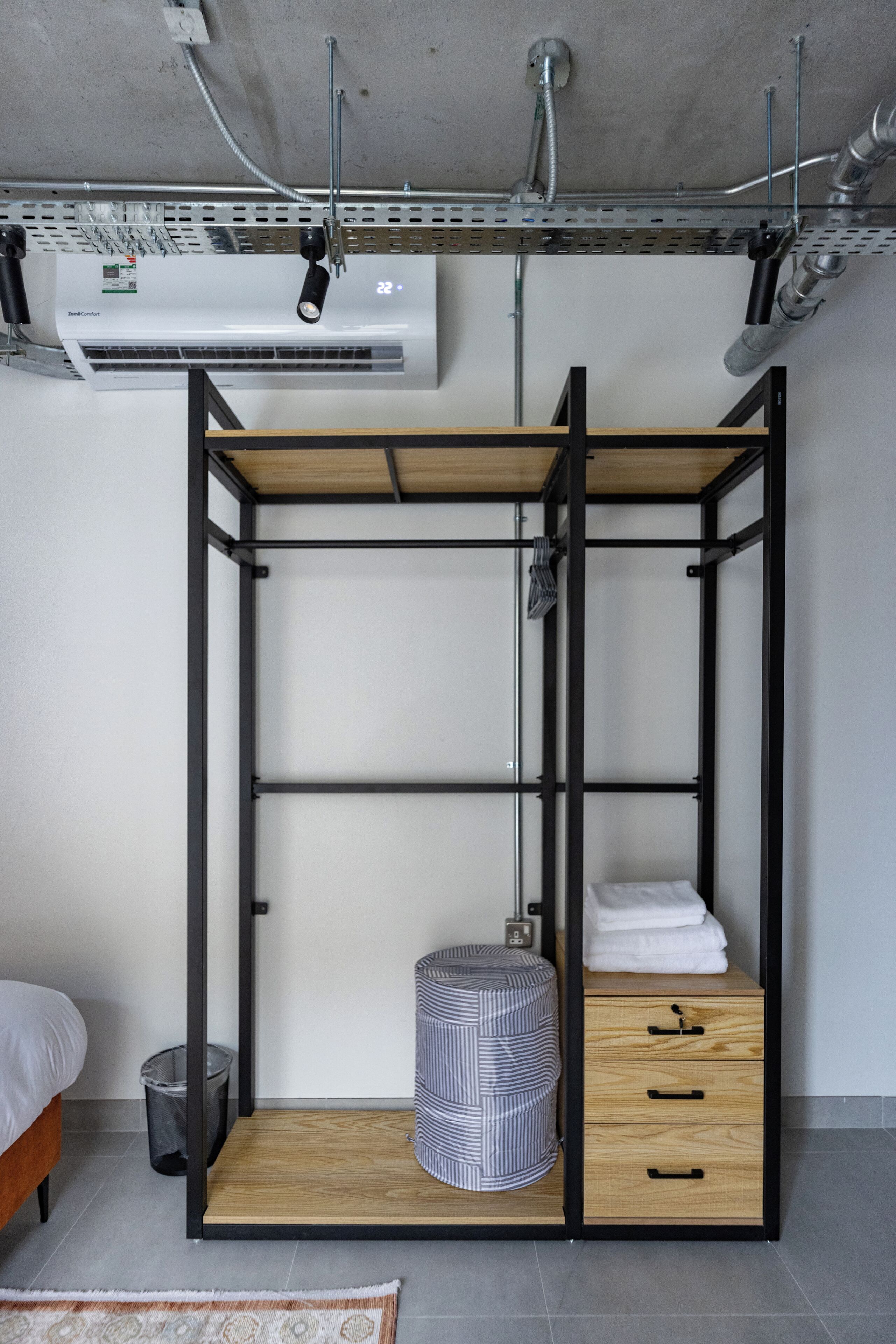 A compact room with a bunk bed, a small wooden cabinet, a striped ottoman, and a trash bin.