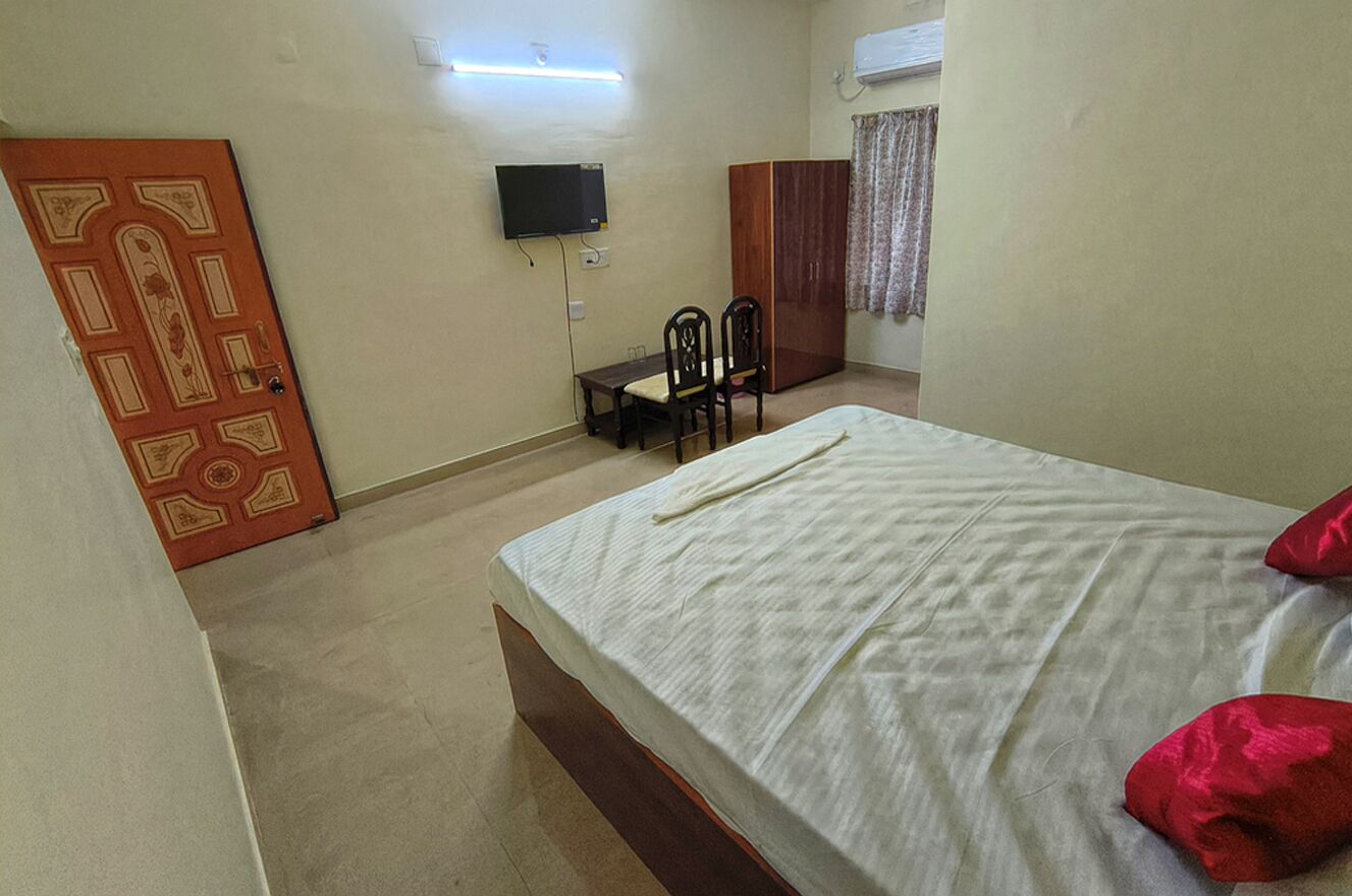 A bedroom with a bed, a TV mounted on the wall, a wardrobe, and a desk with chairs.