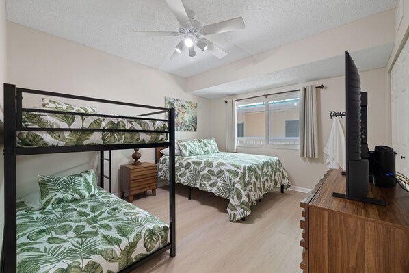 A dormitory room with bunk beds, a ceiling fan, a window with curtains, and a desk with a computer.