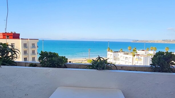 Eine Terrasse mit weißem Tisch und zwei Stühlen, mit Blick auf einen Strand und Gebäude im Hintergrund.