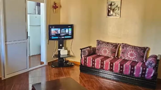 A living room with a dark wooden coffee table, a sofa with patterned cushions, a television, and a chandelier. - Brise d’Océan Tanger (Tangier)