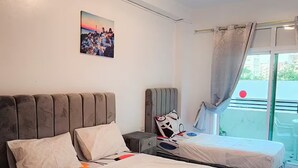 A hotel room with two beds, a bedside table, a chair, a window with curtains, and a wall-mounted picture.