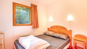 A bedroom with a wooden bed, a window with curtains, a bedside table, and a wall-mounted light. - Charming apartment, sleeps 6, balcony (Modane)