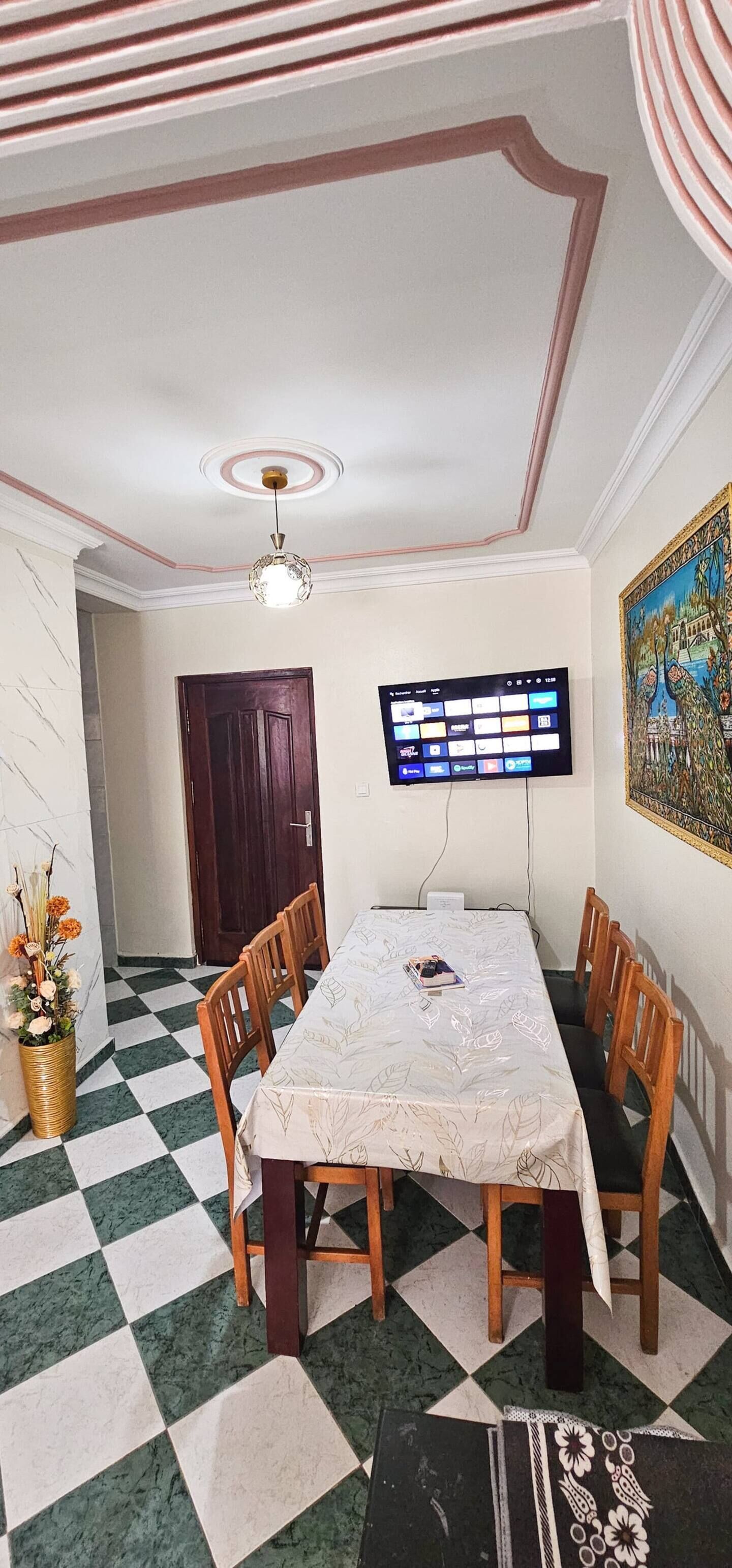 A dining area with a checkered floor, a table with chairs, a ceiling fan, a mounted TV, and a door in the background.