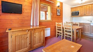 Un intérieur en bois avec un téléviseur fixé au mur, une fenêtre avec des rideaux, un coin cuisine avec des placards en bois et un four à micro-ondes, et une petite table.