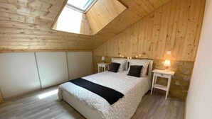 Un dormitorio con techo inclinado, paredes de madera, una cama con colcha blanco y negro, dos mesitas de noche con lámparas y un tragaluz.