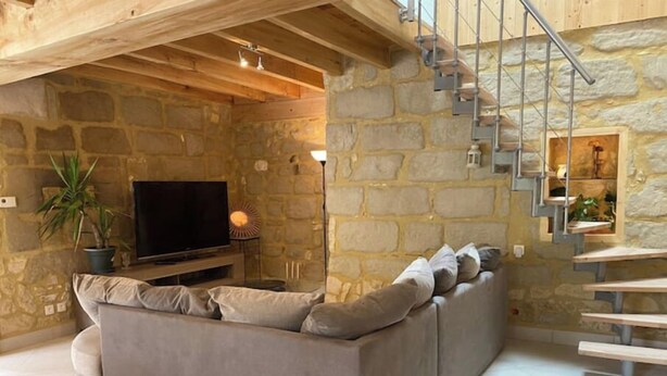 Una sala de estar acogedora con un sofá gris, un televisor de pantalla plana, una planta en maceta y una escalera con un muro de piedra de fondo.