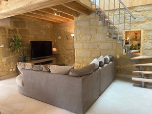 A cozy living room with a grey sofa, a flat-screen TV, a potted plant, and a staircase with a stone wall backdrop. - Gîte (with jacuzzi) Lamothe-d'Alès. Lot et Garonne (Lavergne)
