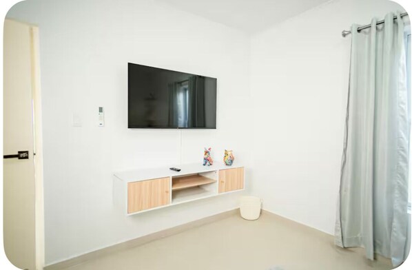 A room with a mounted TV, a white cabinet with wooden doors, a small white bucket, and a light-colored curtain.