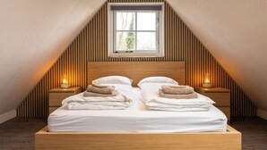 Un dormitorio con techo inclinado, una ventana, dos mesitas de noche con lámparas, una cama con ropa de cama y toallas blancas, y paneles de madera en las paredes.