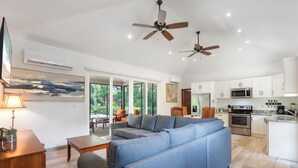 A spacious living area with a grey sectional sofa, wooden coffee table, and a dining table with chairs. There are ceiling fans, a kitchen with white cabinets and stainless steel appliances, and a large window offering a view of greenery.