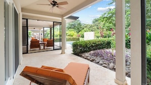 A covered outdoor area with a ceiling fan, a seating area with orange cushions, and a view of a garden with various plants and a stone pathway.