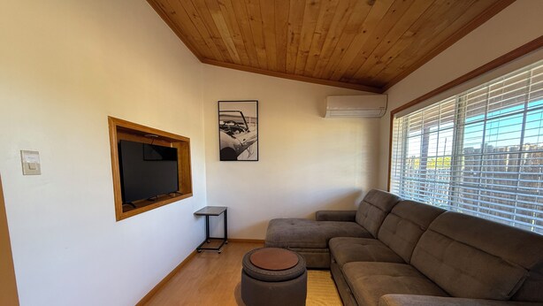 A living room with a wooden ceiling, a large flat-screen TV mounted on the wall, a gray sectional sofa, a small round ottoman, and a window with blinds. - Charming 2-bedroom Guest house in Santa Fe with AC (Santa Fe)