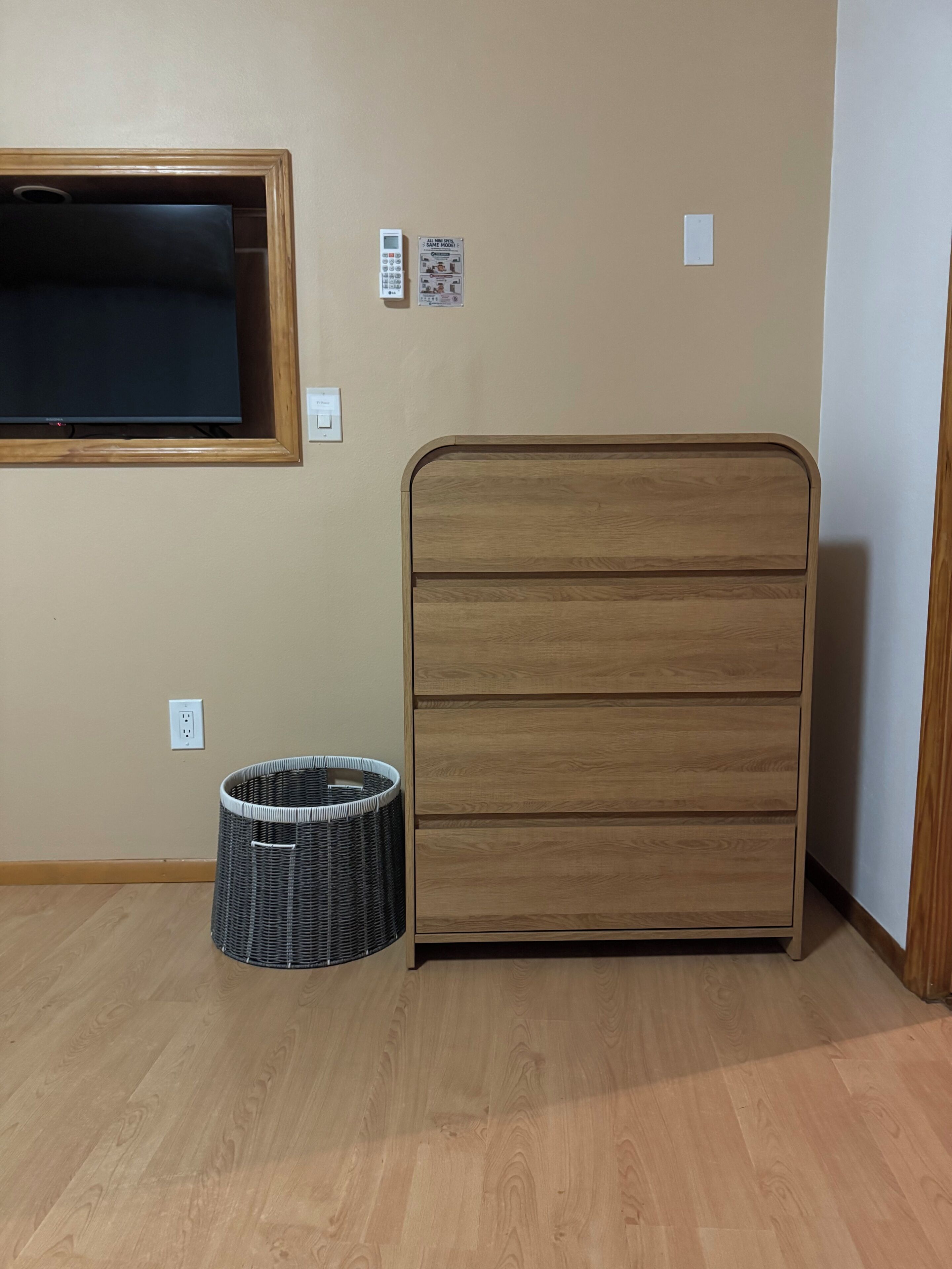 Una habitación con una cama de madera, un televisor montado en la pared, una cesta pequeña y un enchufe.